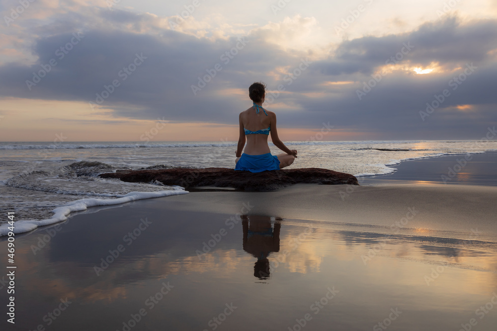 Sunset yoga. Caucasian woman sitting on the stone in Lotus pose. Padmasana. Hands in gyan mudra. Beach in Bali. View from back. Copy space. Yoga retreat. Water reflection. Self care. Zen life.