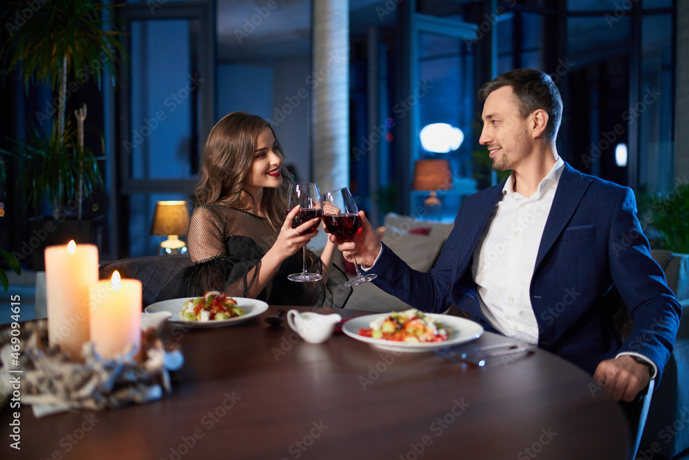 Adorable woman and handsome man in stylish outfits sitting at table, drinking red wine and eating fresh salads. Beautiful couple having romantic dinner at modern apartment with panoramic windows.