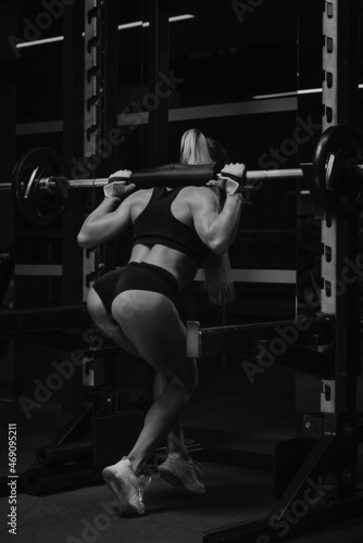 A fit woman with blonde hair is lowering the barbell after squats in a gym.