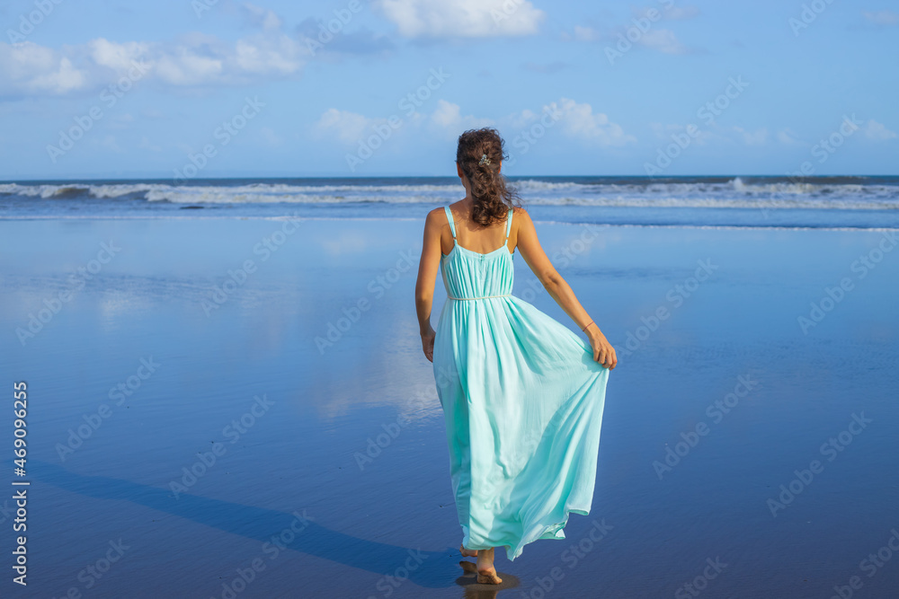 Young woman walking barefoot on empty beach. Full body portrait. Slim Caucasian woman wearing long dress. View from back. Summer sunlight. Blue sky. Travel concept. Bali, Indonesia Young woman walking barefoot on empty beach. Full body portrait. Slim Caucasian woman wearing long dress. View from back. Summer sunlight. Blue sky. Travel concept. Bali, Indonesia