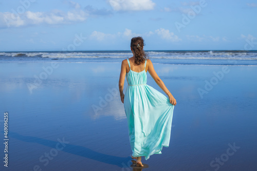 Young woman walking barefoot on empty beach. Full body portrait. Slim Caucasian woman wearing long dress. View from back. Summer sunlight. Blue sky. Travel concept. Bali  Indonesia