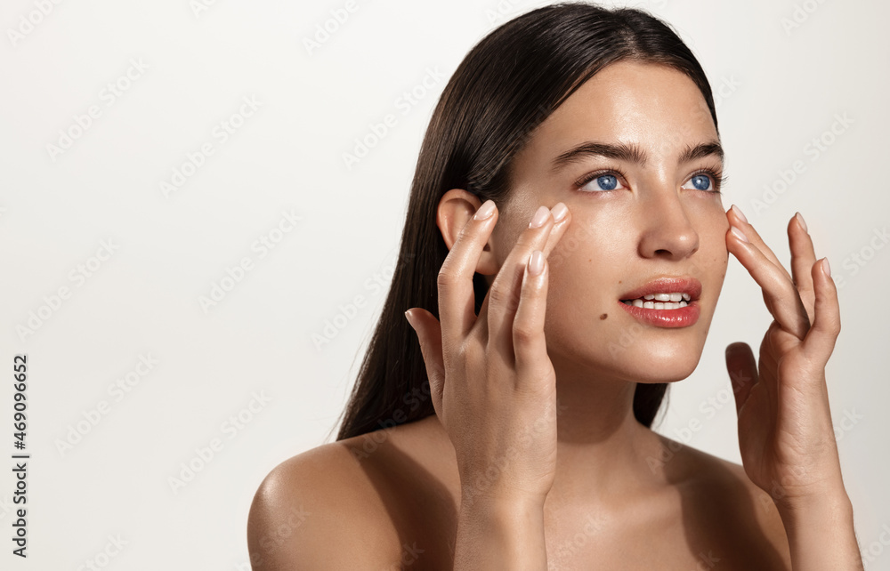 Skin care and women cosmetology. Woman rubbing cream, skincare beauty product on her face, looking away while filling under eye skin with hydrating and nourishing cosmetic products, white background