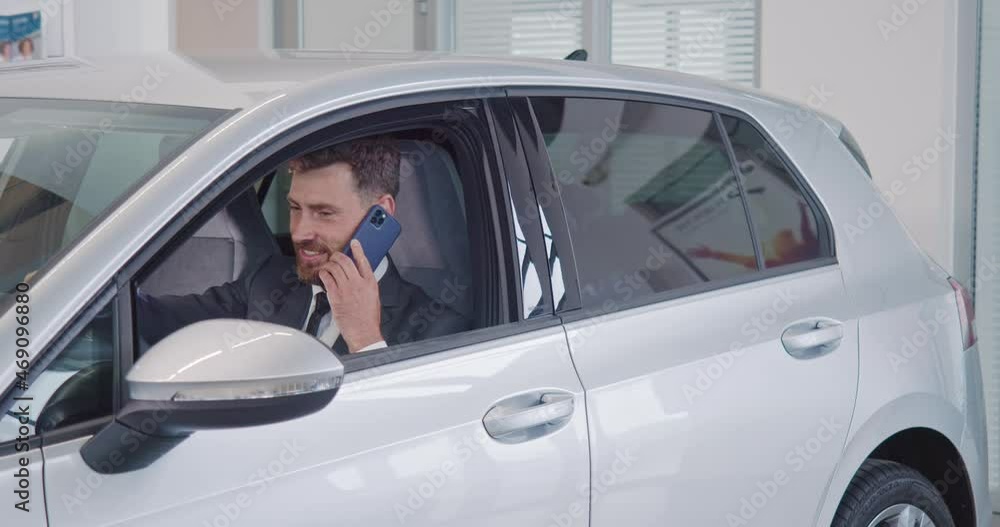 Successful businessman sitting inside new luxury car while talking on smartphone. Bearded man using mobile for consultation about expensive purchase at modern salon. Successful businessman sitting inside new luxury car while talking on smartphone. Bearded man using mobile for consultation about expensive purchase at modern salon.