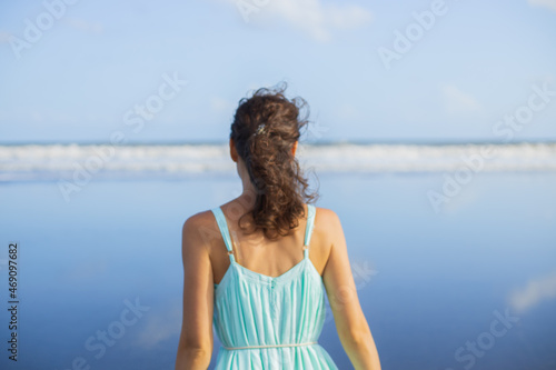 Blurred back view portrait of beautiful Caucasian woman on the beach. Summer vacation in Asia. Young woman wearing dress. Blue sky. Ocean with horizon line. Travel lifestyle. Bali