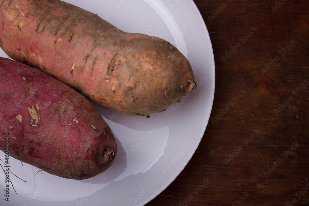 Orange, purple raw sweet potatoes, yam on white plate, wooden rustic table. Farm harvest, organic vegetables, shop local vegan food Orange, purple raw sweet potatoes, yam on white plate, wooden rustic table. Farm harvest, organic vegetables, shop local vegan food