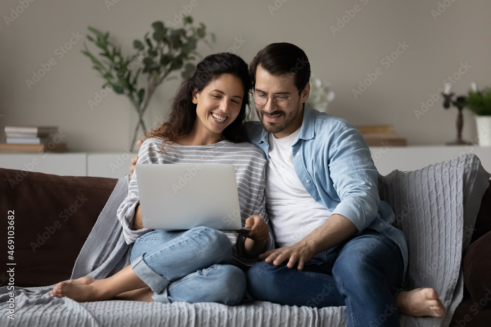 Happy young couple using laptop, relaxing on cozy couch at home together, smiling beautiful woman and man in glasses looking at computer screen, chatting online by video call or watching movie Happy young couple using laptop, relaxing on cozy couch at home together, smiling beautiful woman and man in glasses looking at computer screen, chatting online by video call or watching movie