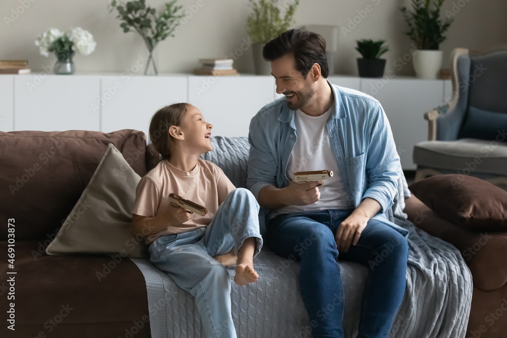 Laughing father and little daughter playing with wooden toy pistols, sitting on comfortable couch at home, smiling dad with 8s girl child engaged in funny activity, spending leisure time together Laughing father and little daughter playing with wooden toy pistols, sitting on comfortable couch at home, smiling dad with 8s girl child engaged in funny activity, spending leisure time together