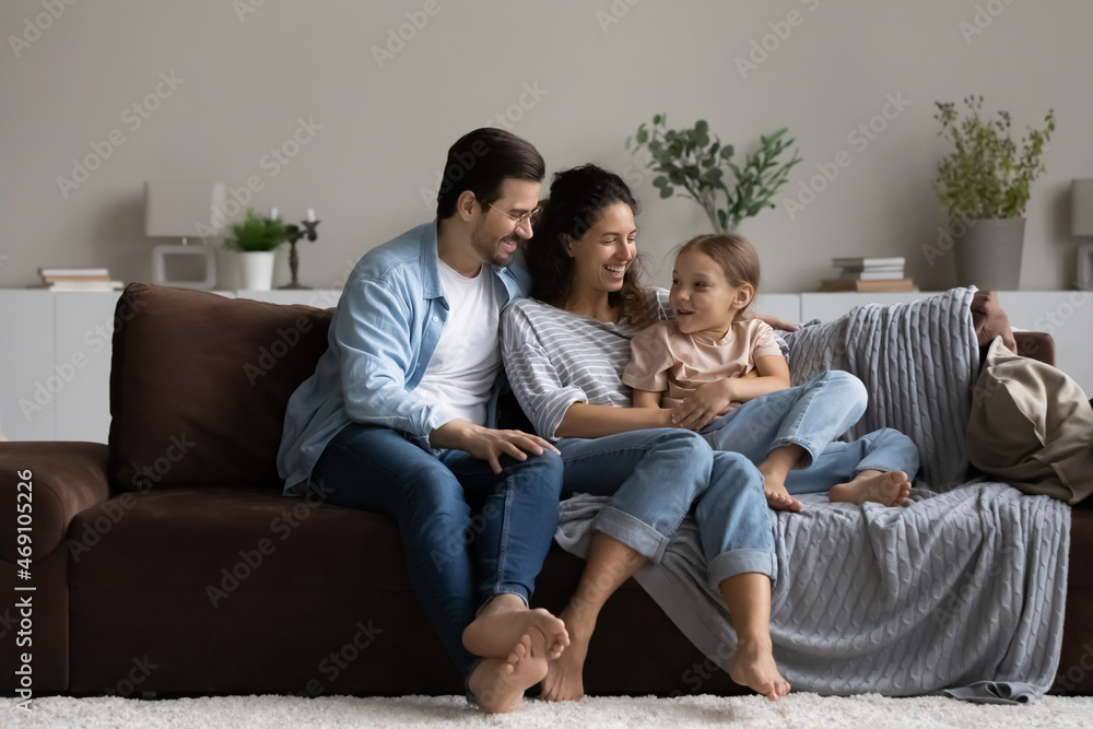 Happy parents with 8s daughter relaxing on comfortable couch together, laughing mother and smiling father in glasses with adorable girl kid having fun, chatting, spending leisure time at home