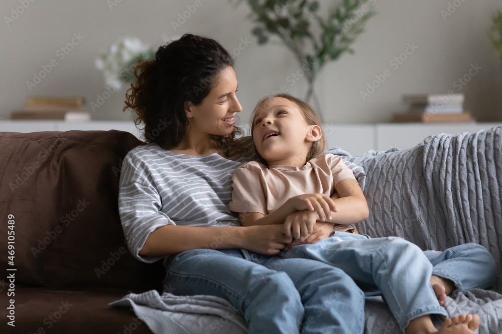 Happy mother with daughter hugging, enjoying tender moment, relaxing on couch at home, happy little girl sitting on loving young mom laps, talking and having fun, spending leisure time together Happy mother with daughter hugging, enjoying tender moment, relaxing on couch at home, happy little girl sitting on loving young mom laps, talking and having fun, spending leisure time together