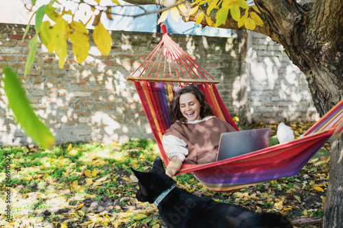 Young happy woman freelancer with laptop and dog in hammock at backyard of country house on autumn day photo