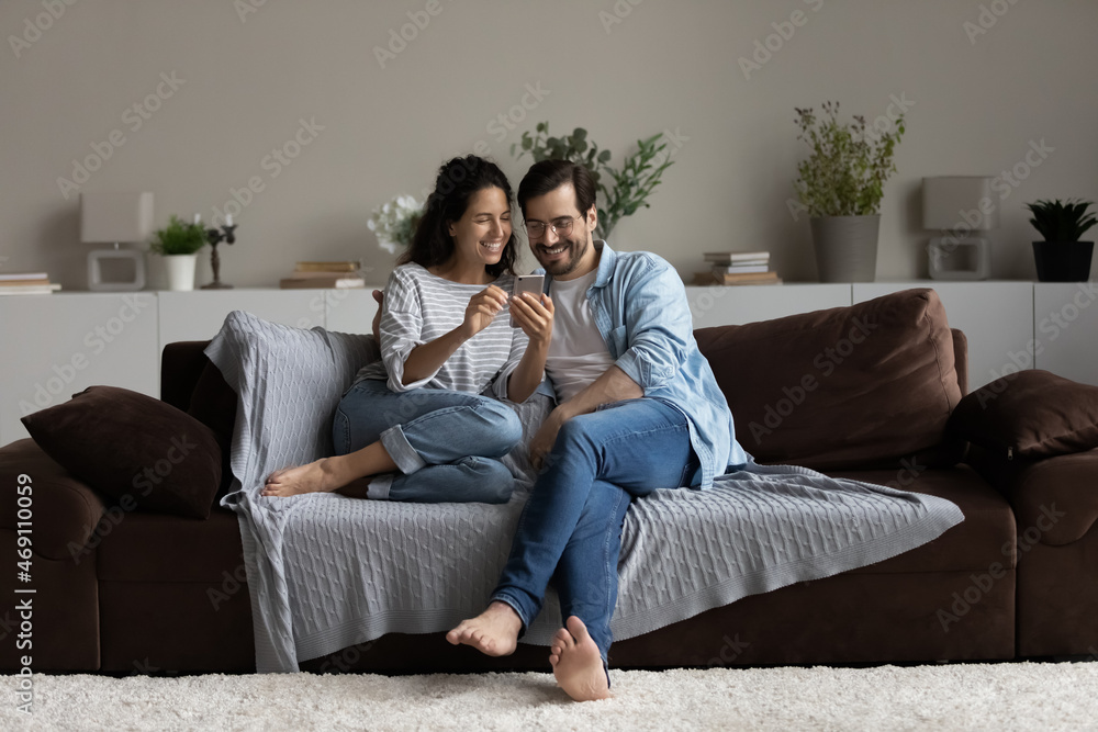 Happy young couple having fun with smartphone at home, sitting on cozy couch, smiling beautiful woman and man in glasses looking at phone screen, watching funny video, surfing internet, chatting Happy young couple having fun with smartphone at home, sitting on cozy couch, smiling beautiful woman and man in glasses looking at phone screen, watching funny video, surfing internet, chatting