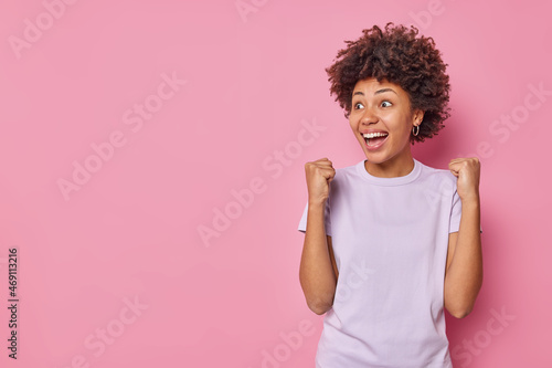 Relieved positive young woman with curly hair triumphs as celebrates success clenches fists watches sports game stares with eyes full of happiness dressed casually isolated over pink background