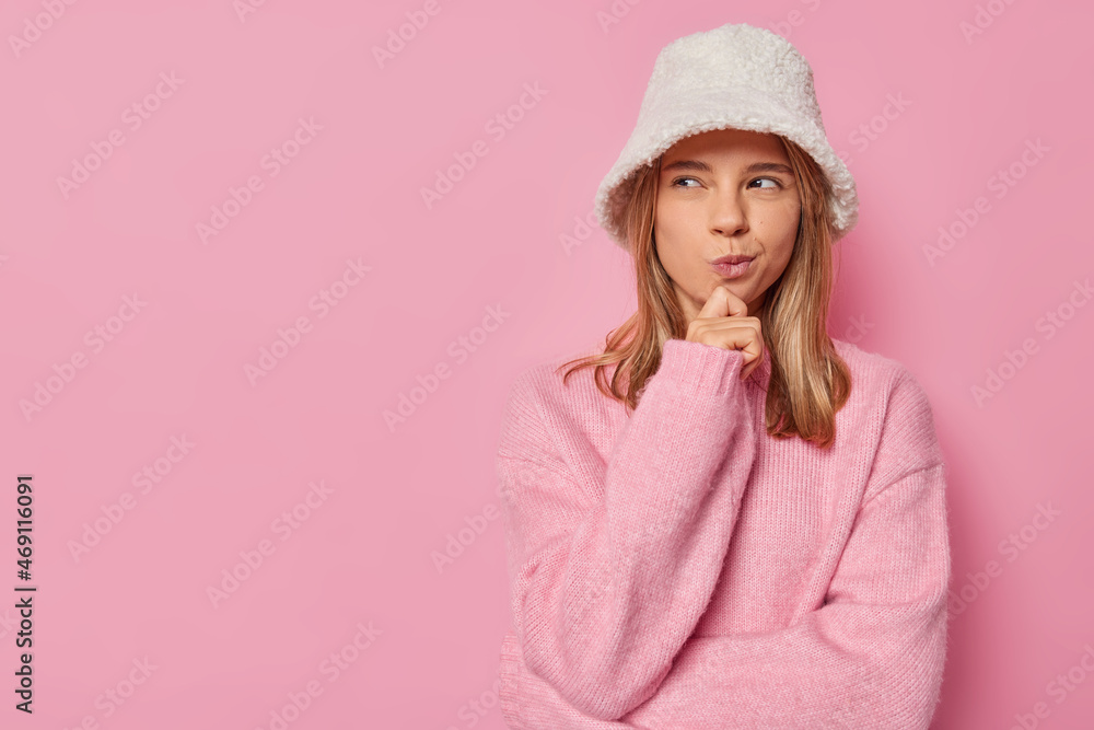 Obraz premium Horizontal shot of lovely pensive woman holds chin purses lips and looks mysteriously away wears white stylish hat and casual loose jumper isolated over pink background with copy space on right
