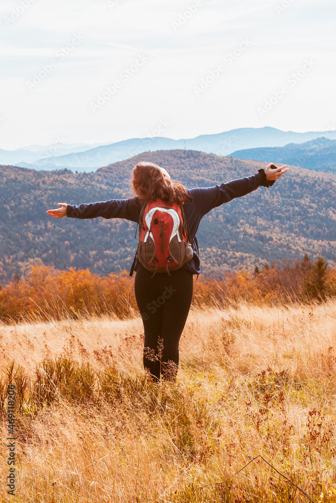 Fototapeta premium A girl with a backpack in the tall dry grass enjoys the autumn view of the mountains