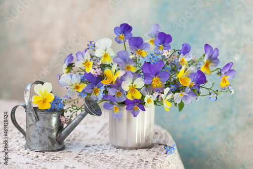 Fényképezés A bouquet of pansies and forget-me-not flowers in a vase and a watering can on a table with a tablecloth against the background of a colored wall