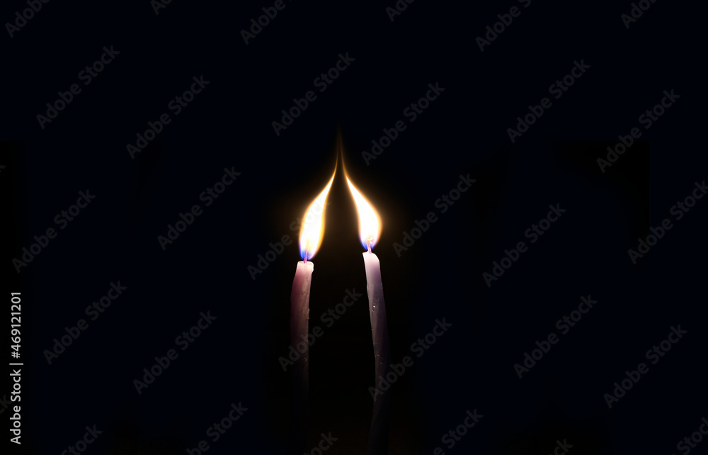 Two thin candles stretch their lights towards each other. The candles are in complete darkness. Black background. Two thin candles stretch their lights towards each other. The candles are in complete darkness. Black background.