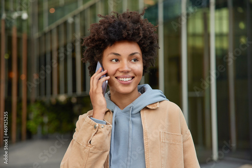 Modern technologies and people concept. Happy young curly haired woman enjoys cellular communication smiles positively dressed casually poses outdoors against blurred background makes telephone call