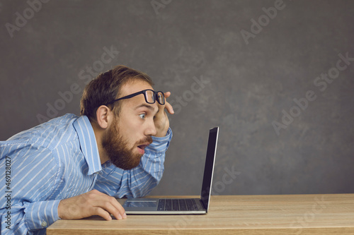 Quadro su tela Side view of a man with a surprised expression sitting at a table and staring at a laptop screen