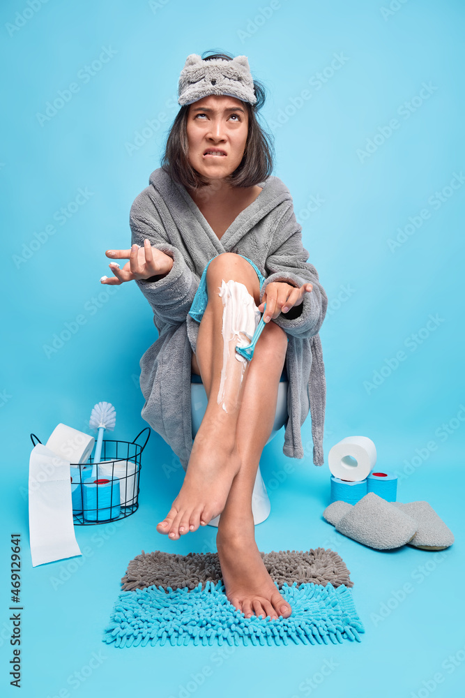 Upset young woman shaves legs with razor being in hurry for date sits on toilet bowl in lavatory room wears grey soft bathrobe and sleepmask isolated over blue background with paper rollers around Upset young woman shaves legs with razor being in hurry for date sits on toilet bowl in lavatory room wears grey soft bathrobe and sleepmask isolated over blue background with paper rollers around