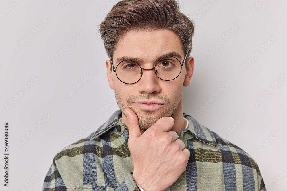 Close up shot of self confident man holds chin looks attentively at camera considers something wears round spectacles and checkered shirt poses against white background. Thoughtful handsome guy Close up shot of self confident man holds chin looks attentively at camera considers something wears round spectacles and checkered shirt poses against white background. Thoughtful handsome guy