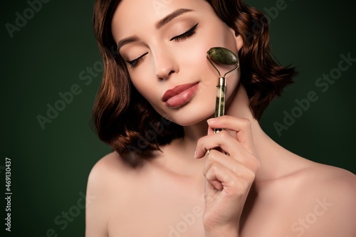 Photo of calm peaceful attractive woman using derma roller on cheek pampering isolated on green color background photo