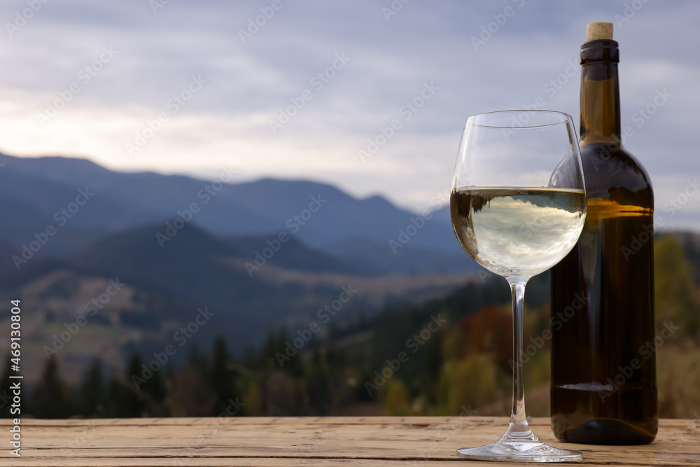 Glass and bottle of tasty wine on wooden table against mountain landscape. Space for text