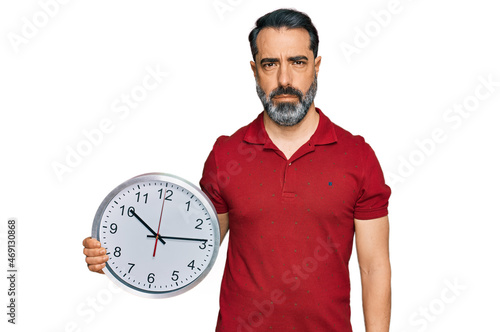 Middle aged man with beard holding big clock thinking attitude and sober expression looking self confident