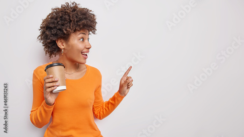 Happy surprised young woman sees something awesome and unexpected indicates at empty blank space away shows place for your advertisement drinks coffee from disposabe cup wears orange jumper.