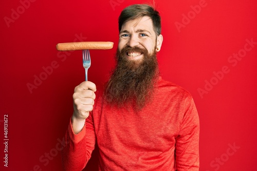 Redhead man with long beard holding fork with pork sausage looking positive and happy standing and smiling with a confident smile showing teeth