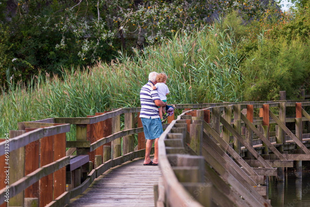 Nonno con nipote in braccio passeggia su un piccolo ponte nella riserva naturale nella laguna del mare adriatico a marano lagunare. Passeggiata estiva tra la natura.