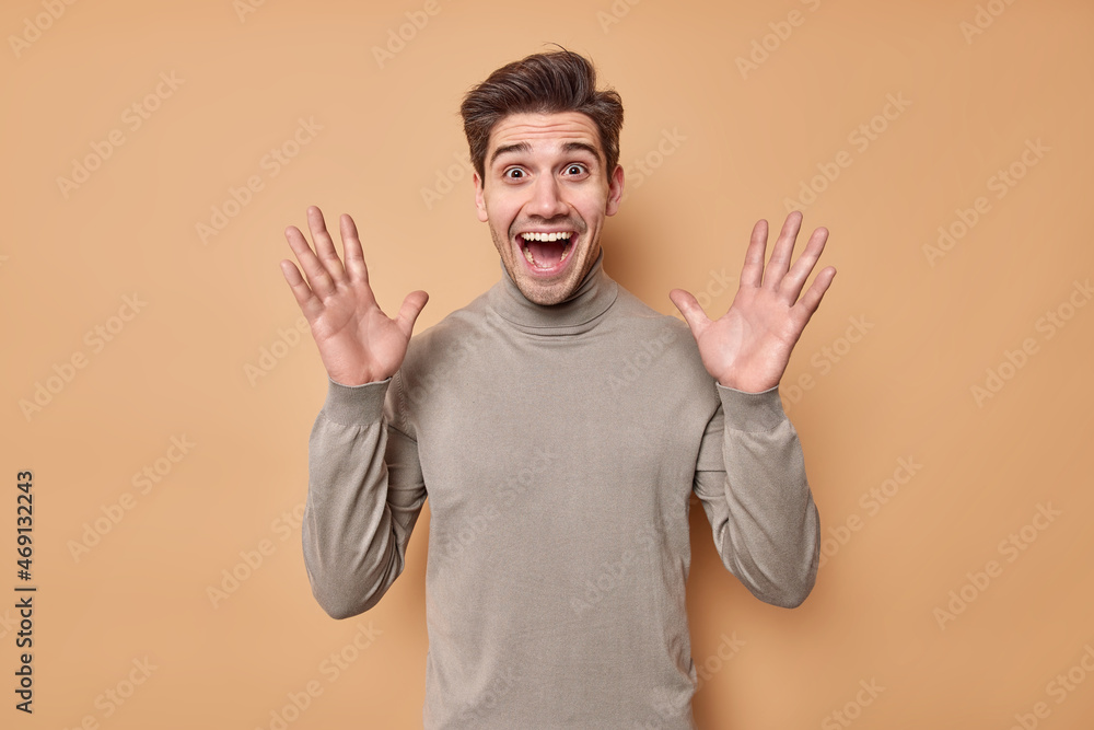 Waist up shot of emotional excited man raises palms feels joyful reacts on awesome nes keeps mouth opened dressed in casual turtleneck isolated over beige backgound. Human reactions concept. Waist up shot of emotional excited man raises palms feels joyful reacts on awesome nes keeps mouth opened dressed in casual turtleneck isolated over beige backgound. Human reactions concept.