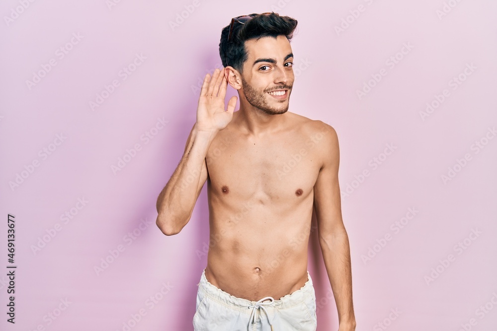 Fototapeta premium Young hispanic man wearing swimwear shirtless smiling with hand over ear listening an hearing to rumor or gossip. deafness concept.