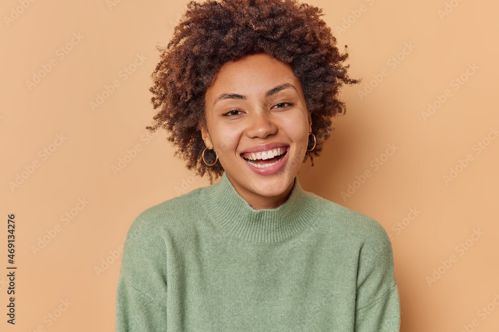 Portrait of cheerful woman with curly hair toothy smile has carefree happy expression wears casual jumper poses against beige background. Positive human emotions and face expressions concept Portrait of cheerful woman with curly hair toothy smile has carefree happy expression wears casual jumper poses against beige background. Positive human emotions and face expressions concept