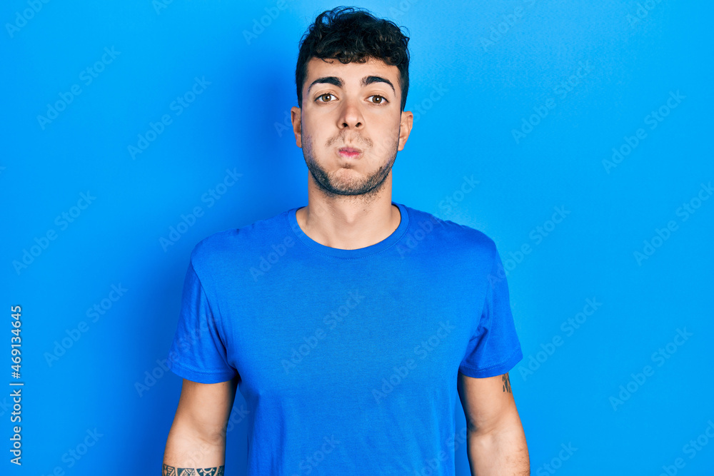 Fototapeta premium Young hispanic man wearing casual blue t shirt puffing cheeks with funny face. mouth inflated with air, crazy expression.