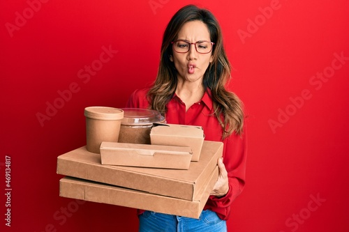 Young latin woman holding take away food making fish face with mouth and squinting eyes, crazy and comical.