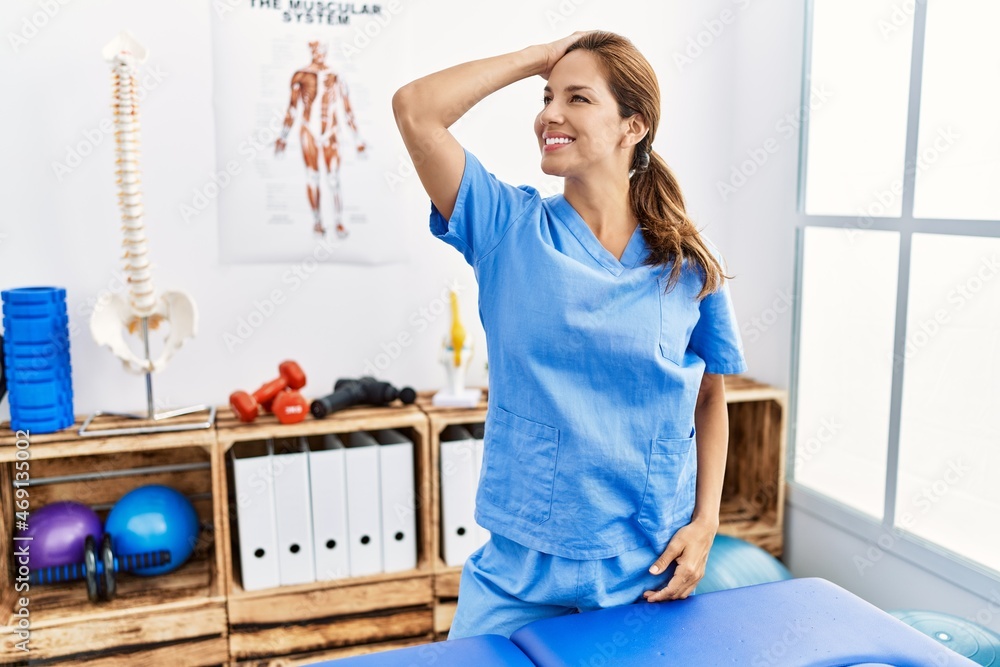 Middle age hispanic physiotherapist woman working at pain recovery clinic smiling confident touching hair with hand up gesture, posing attractive and fashionable Middle age hispanic physiotherapist woman working at pain recovery clinic smiling confident touching hair with hand up gesture, posing attractive and fashionable