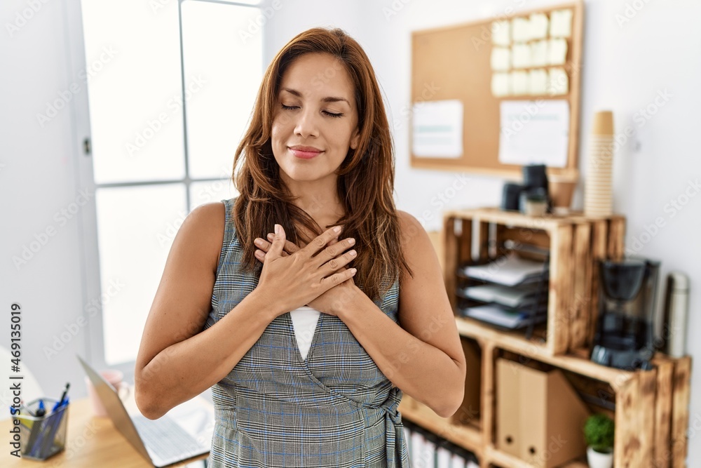 Middle age hispanic woman at the office smiling with hands on chest with closed eyes and grateful gesture on face. health concept. Middle age hispanic woman at the office smiling with hands on chest with closed eyes and grateful gesture on face. health concept.