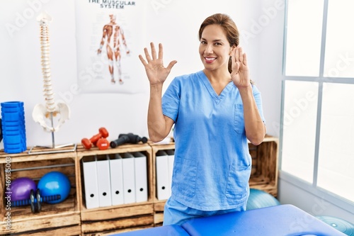 Middle age hispanic physiotherapist woman working at pain recovery clinic showing and pointing up with fingers number nine while smiling confident and happy.