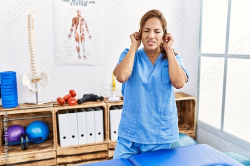 Middle age hispanic physiotherapist woman working at pain recovery clinic covering ears with fingers with annoyed expression for the noise of loud music. deaf concept.