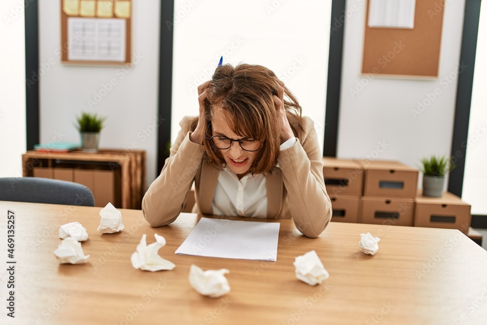 Young caucasian businesswoman stressed with hands on head writing on paperwork at the office.