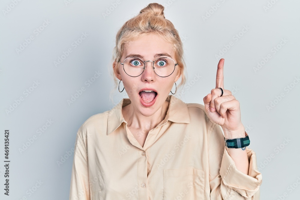 Beautiful caucasian woman with blond hair wearing casual look and glasses pointing finger up with successful idea. exited and happy. number one.