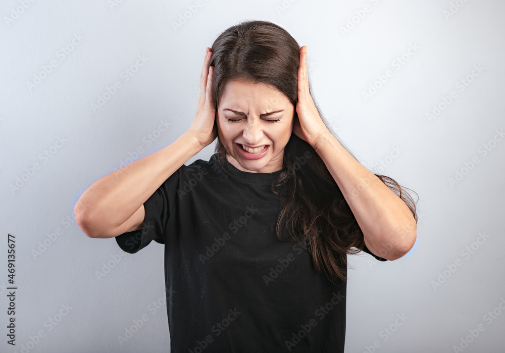 Unhappy stressed grimacing frowns woman not want to listen and covering the ears her fingers on blue background. Closeup Unhappy stressed grimacing frowns woman not want to listen and covering the ears her fingers on blue background. Closeup