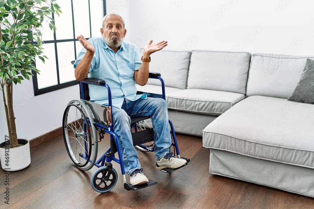 Handsome senior man sitting on wheelchair at the living room clueless and confused expression with arms and hands raised. doubt concept. Handsome senior man sitting on wheelchair at the living room clueless and confused expression with arms and hands raised. doubt concept.
