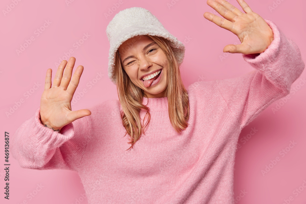 Funny positive European woman tilts head winks eyes and keeps palms raised wears hat and jumper poses against pink background expresses happy feelings feels carefree. Human emotions concept.