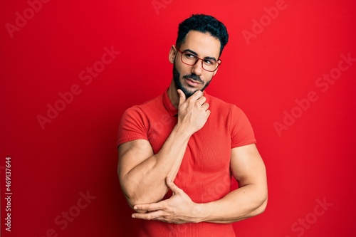 Young hispanic man wearing casual clothes and glasses with hand on chin thinking about question, pensive expression. smiling with thoughtful face. doubt concept.