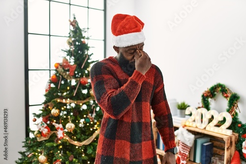 African american man wearing santa claus hat standing by christmas tree tired rubbing nose and eyes feeling fatigue and headache. stress and frustration concept.