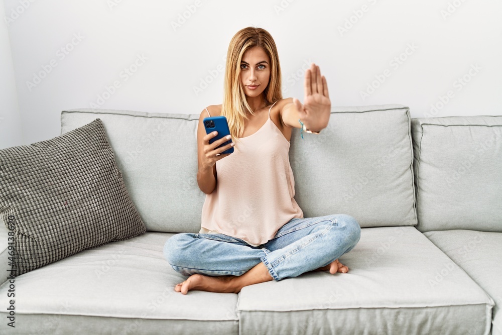 Blonde beautiful young woman sitting on the sofa at home using smartphone doing stop sing with palm of the hand. warning expression with negative and serious gesture on the face. Blonde beautiful young woman sitting on the sofa at home using smartphone doing stop sing with palm of the hand. warning expression with negative and serious gesture on the face.