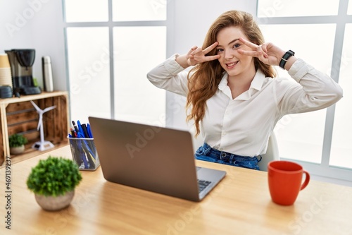 Young caucasian woman working at the office using computer laptop doing peace symbol with fingers over face, smiling cheerful showing victory