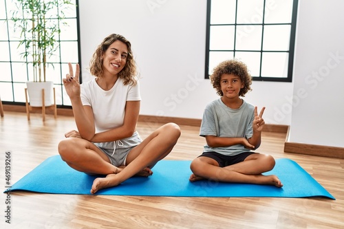 Young woman and son sitting on training mat at the gym smiling with happy face winking at the camera doing victory sign. number two.
