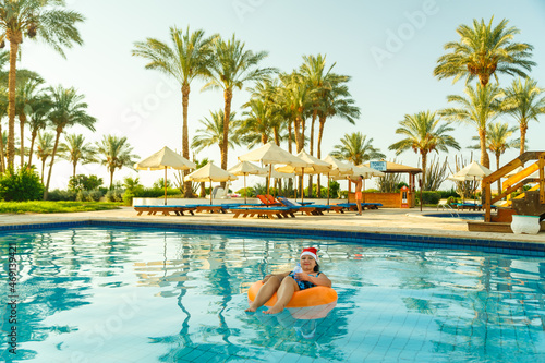 joyful woman in a santa claus cap in a swimming circle in the pool with a cocktail in hand in the new year holidays on the background of palm trees. © finist_4